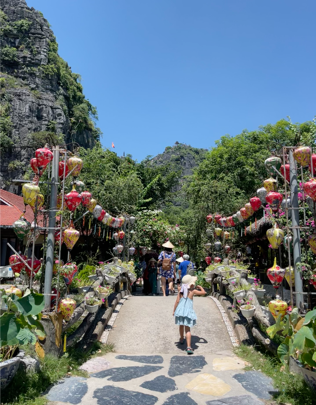 a child in a sunhat walks down a path lined with bright colored lanterns and with a view of large karsts in the background.